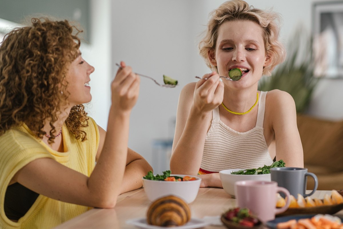 Two smiling women sharing a salad at a table with fruit and pastries