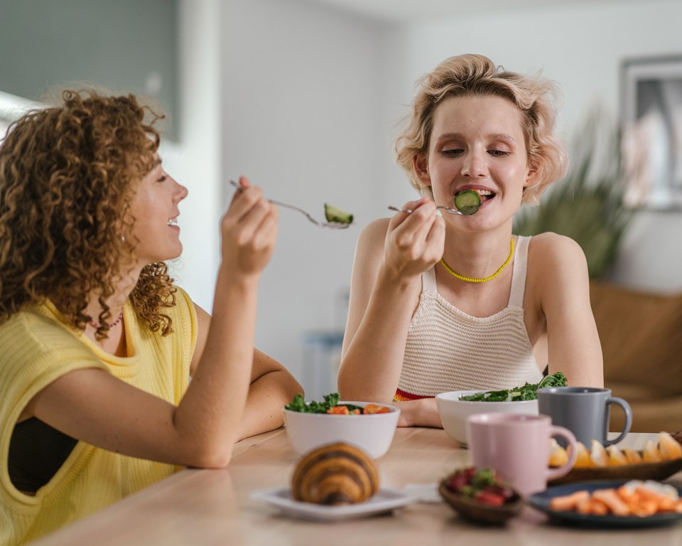 Two smiling women sharing a salad at a table with fruit and pastries