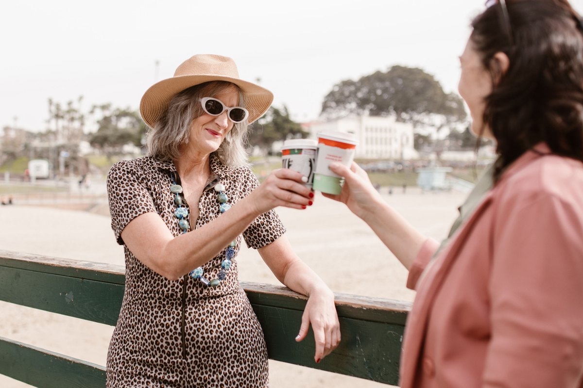 Two older women clinking takeaway coffee cups at a railing on a beach boardwalk
